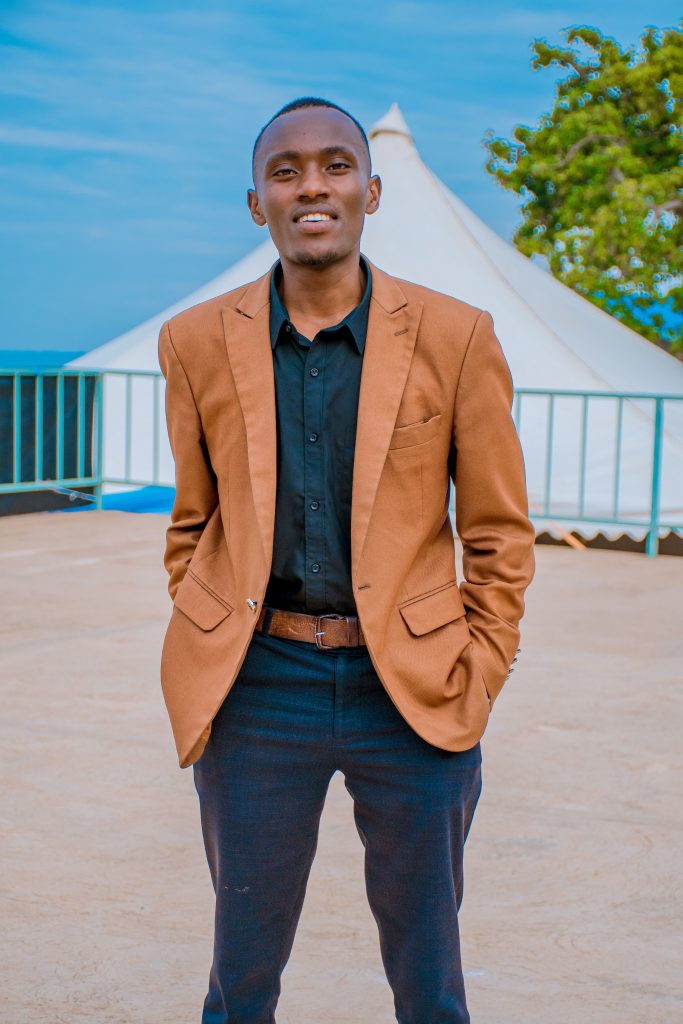 Confident man in a brown blazer standing outdoors with a tent in the background.